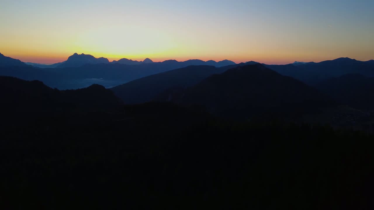 Standbild aus dem Drohnenflug: gestaffelte Bergkämme als Silhouette vor einem orange leuchtenden Dämmerungshimmel